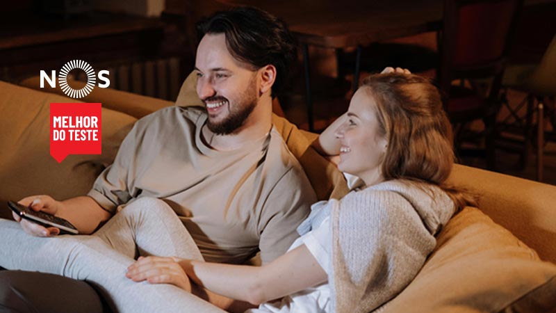 Casal feliz, num sofá, a ver televisão com o pacote de telecomunicações NOS.